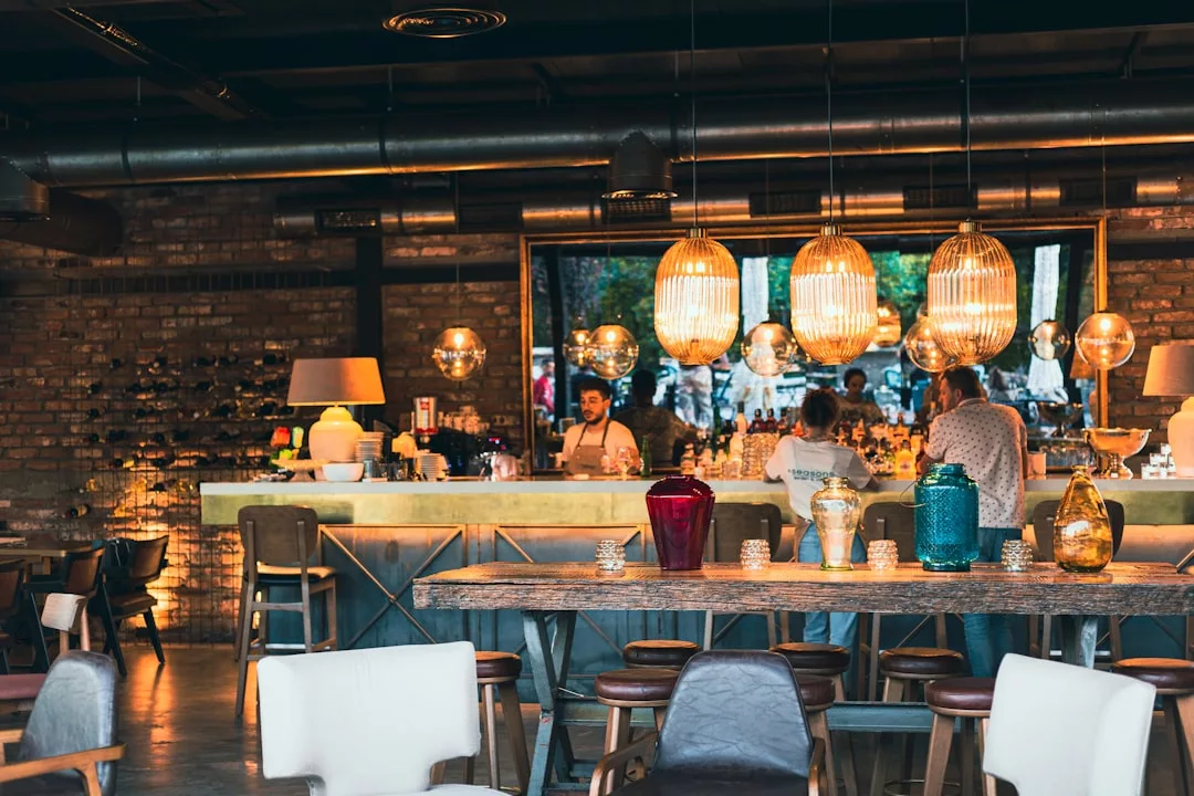 Moderne Mallorca-Bar mit Hängelampen, Barhockern und Gästen am Tresen; freiliegende Backsteinwände und dekorative Vasen auf einem zentralen Tisch.