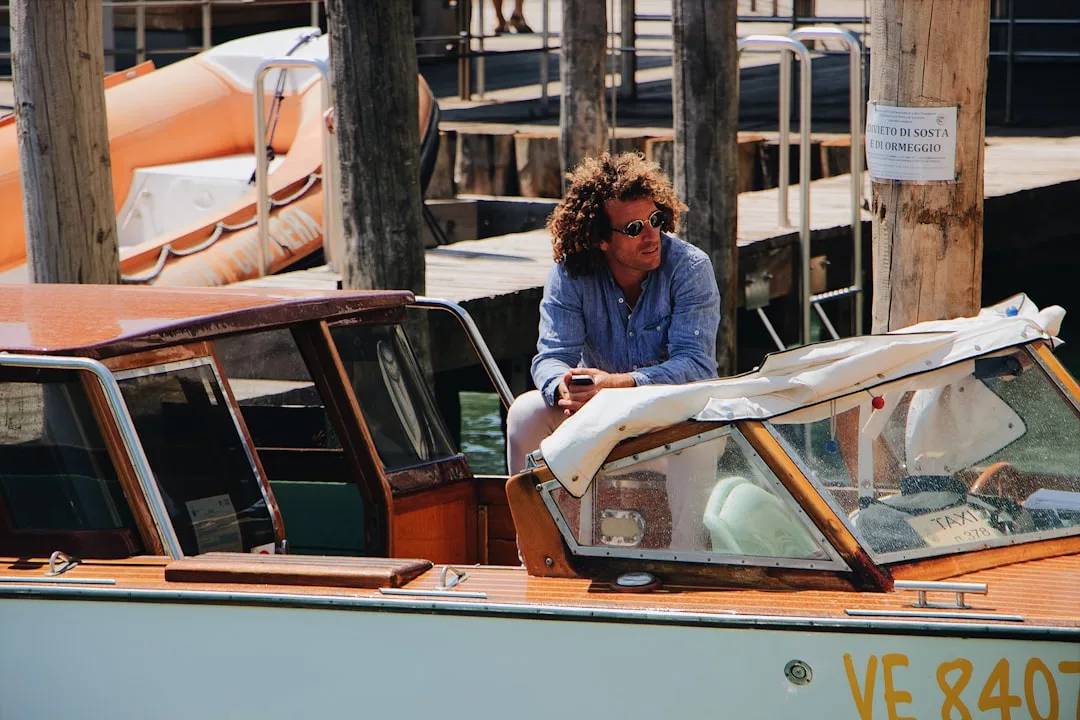 Ein Mann mit lockigem Haar und Sonnenbrille sitzt auf dem Rand eines Holzbootes, das in einem Jachthafen auf Mallorca angedockt ist. Im Hintergrund sind Holzpfähle und ein weiteres Boot zu sehen.