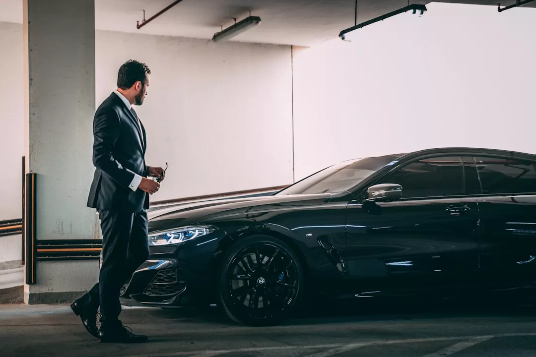 Ein Mann im Anzug steht neben einem schwarzen Luxuswagen in einem Parkhaus auf Mallorca, hält eine Sonnenbrille in der Hand und blickt in Richtung des Fahrzeugs.