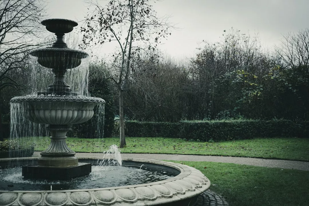 Ein Steinbrunnen mit fließendem Wasser steht in einem Park auf Mallorca, umgeben von grünem Gras, Bäumen ohne Blätter und bedecktem Himmel.