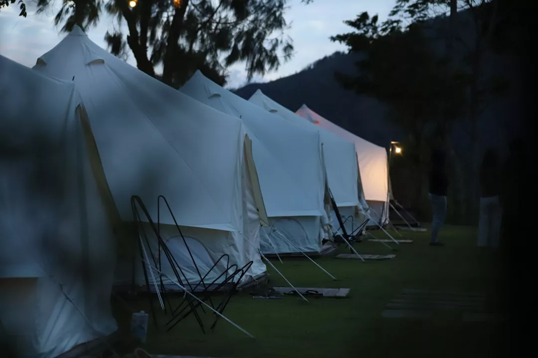 Eine Reihe weißer Zelte, die in der Abenddämmerung auf einer Wiese auf Mallorca aufgebaut sind, mit einer Bergsilhouette und Bäumen im Hintergrund. Einige Zelte sind von innen beleuchtet.