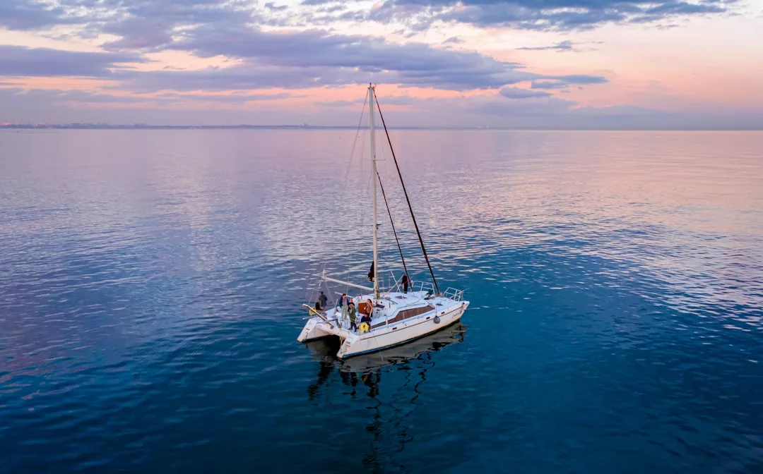 Ein weißes Segelboot mit Menschen an Bord treibt auf ruhigem Wasser vor der Küste Mallorcas unter einem teilweise bewölkten Himmel bei Sonnenuntergang.