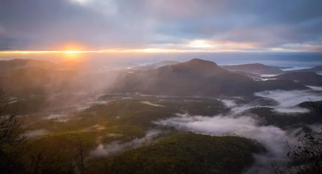Sonnenaufgang über den nebelverhangenen Bergen Mallorcas mit dichten grünen Wäldern und tief hängenden Wolken, unter einem teilweise bewölkten Himmel.