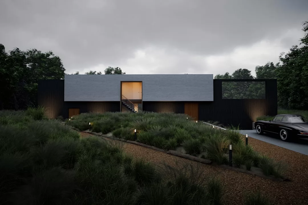 Ein modernes, rechteckiges Haus mit dunklen Außenwänden und großen Fenstern, inspiriert vom mallorquinischen Stil, ist von hohem Gras und einem Kiesweg umgeben, auf dem rechts ein Auto geparkt ist.