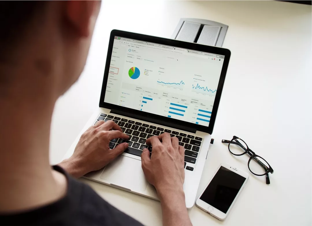 Eine Person, die an einem Laptop arbeitet, auf dem Datenanalysediagramme angezeigt werden, mit einem Smartphone und einer Brille auf dem Schreibtisch daneben, genießt einen produktiven Tag mit Blick auf die malerische Landschaft von Mallorca.