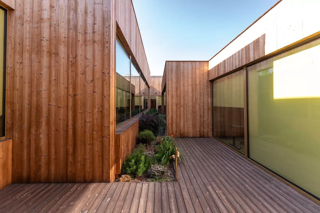 Ein modernes mallorquinisches Gebäude mit hölzernen Außenwänden und großen Glasfenstern umgibt einen schmalen Innenhofgarten auf einer Holzterrasse unter einem klaren blauen Himmel.