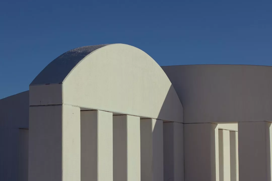 Weiße geometrische Gebäudestruktur mit vertikalen Säulen und einem gewölbten Dachabschnitt vor einem klaren blauen Himmel über Mallorca.
