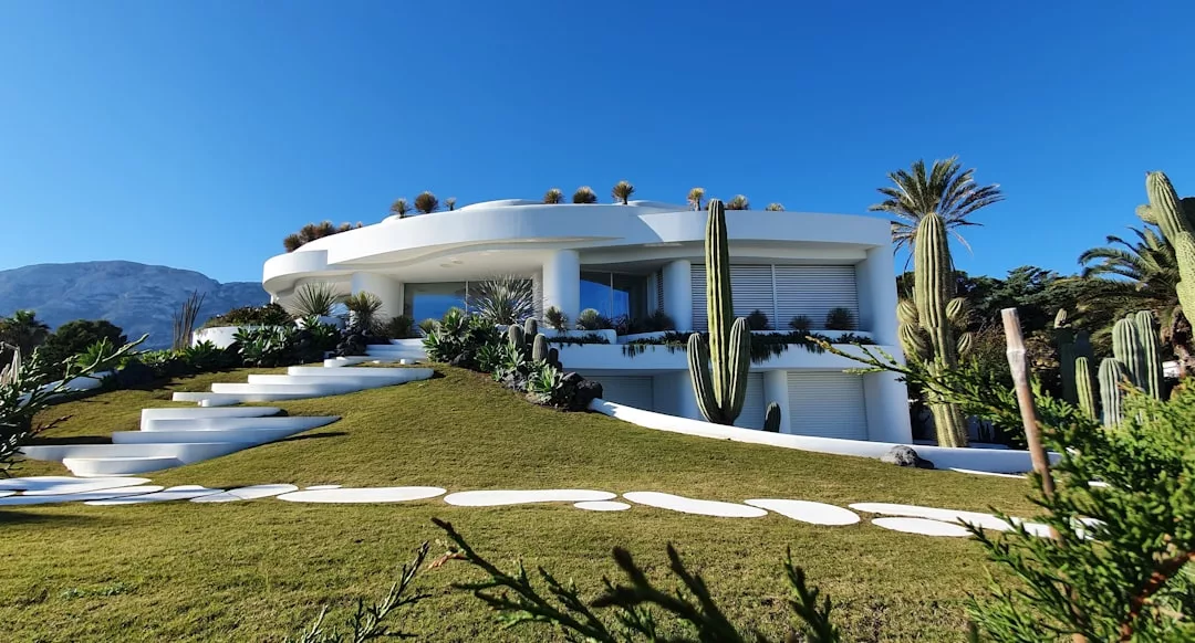 Modernes weißes zweistöckiges Haus mit geschwungener Architektur auf Mallorca, umgeben von Kakteen und anderen Wüstenpflanzen, auf einem grasbewachsenen Hügel unter strahlend blauem Himmel gelegen.