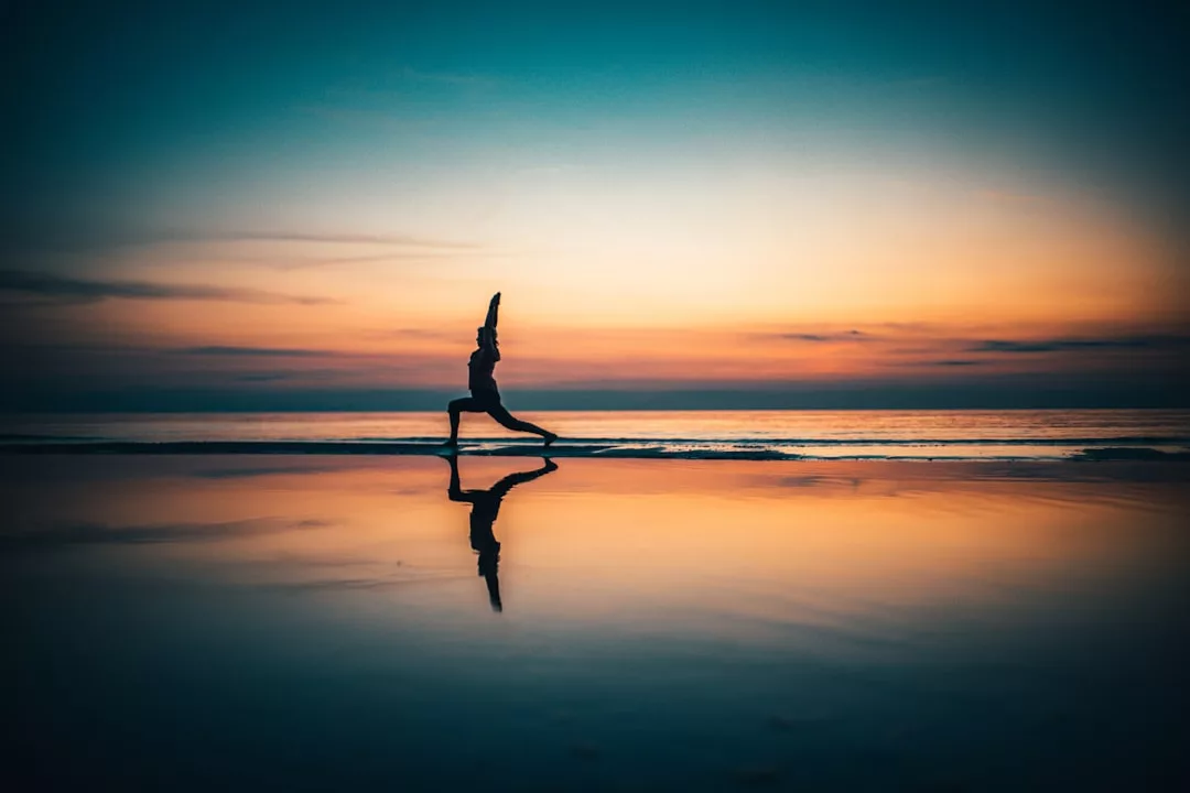 Eine Person übt an einem Strand auf Mallorca bei Sonnenuntergang Yoga im Ausfallschritt, ihr Spiegelbild schimmert im nassen Sand.