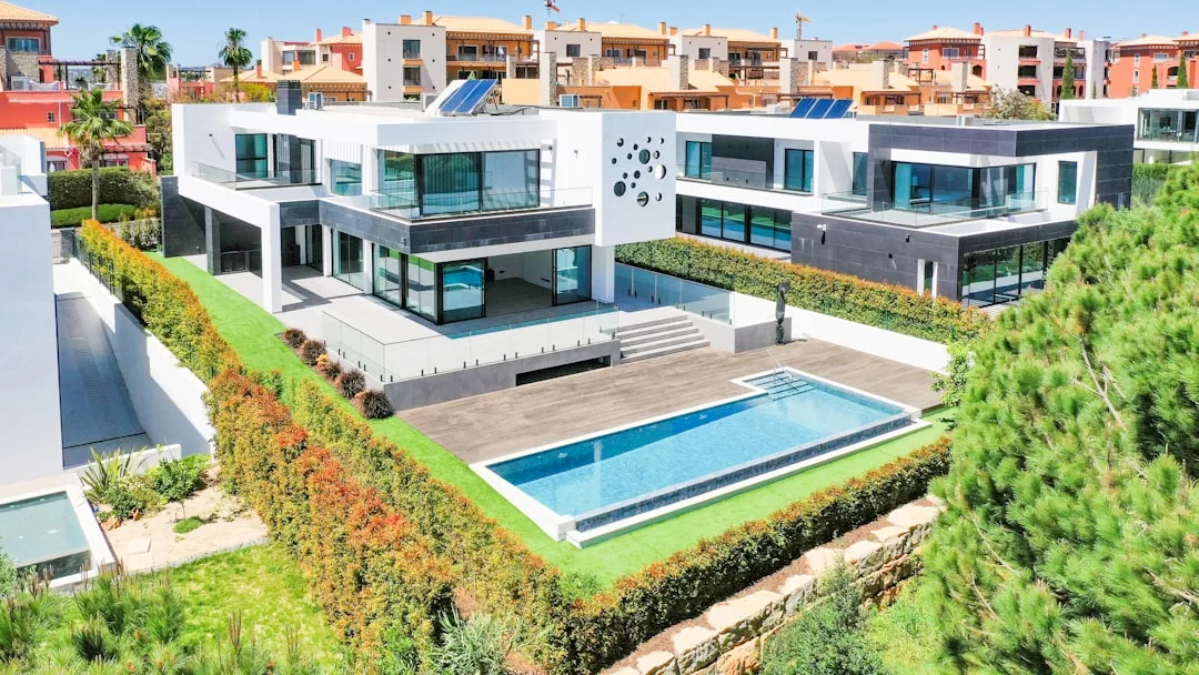 Modernes zweistöckiges Haus auf Mallorca mit großen Glasfenstern, einem Pool und gepflegtem Rasen, umgeben von Hecken; im Hintergrund sind die Nachbargebäude zu sehen.