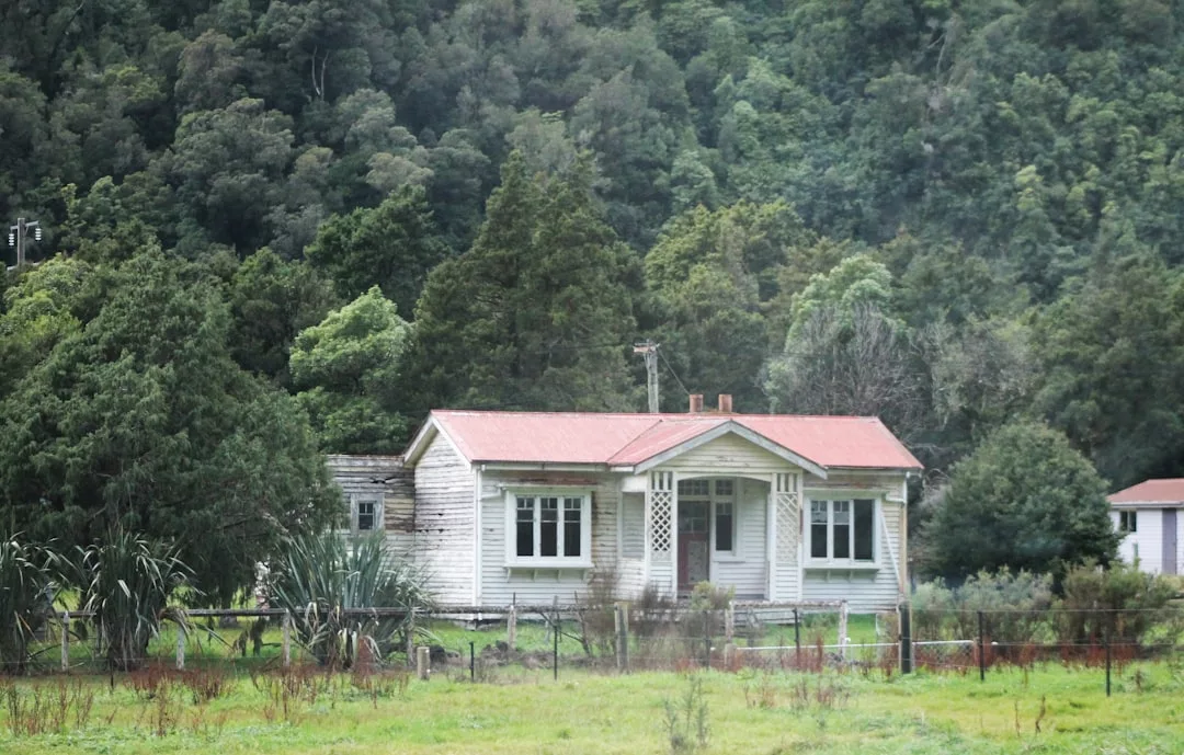Ein kleines, verwittertes Haus mit rotem Dach steht vor einem dichten grünen Wald auf Mallorca, umgeben von Gras und Bäumen, mit einem Zaun im Vordergrund.