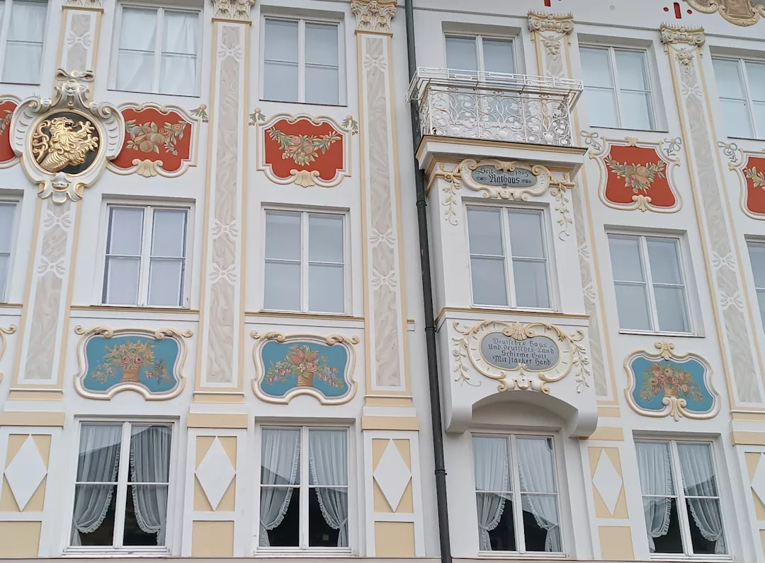 Verschnörkelte mallorquinische Gebäudefassade mit Zierleisten, bunten Paneelen, goldenen Akzenten und mehreren Fenstern mit weißen Vorhängen; ein Teil weist einen kleinen, verzierten Balkon auf.