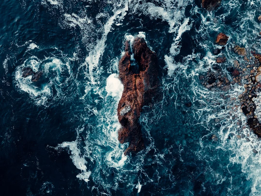 Luftaufnahme von Meereswellen, die gegen einen großen Felsen vor der Küste Mallorcas schlagen, umgeben von wirbelndem blauen Wasser und kleineren Felsen.