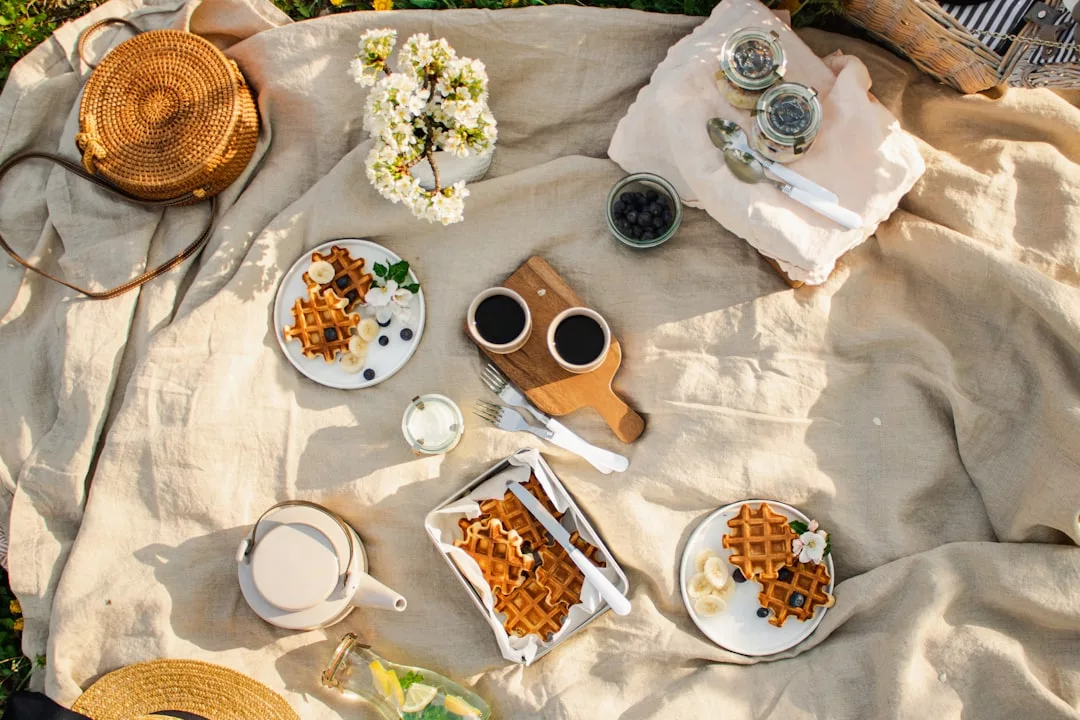 Ein Picknick auf einer beigen Decke mit Tellern mit Waffeln, Blaubeeren, Kaffee, einer Teekanne, Blumen und Körben, die auf dem Gras arrangiert sind, erinnert an den entspannten Charme von Mallorcas sonnenverwöhnten Morgenstunden.