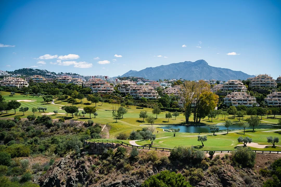 Panoramablick auf einen mallorquinischen Golfplatz mit grünen Fairways, verstreuten Bäumen, einem Teich und Wohngebäuden unter einem klaren blauen Himmel mit Bergen im Hintergrund.