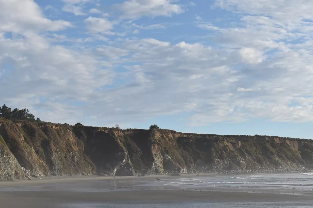 Felsige Klippen säumen einen Sandstrand auf Mallorca bei meist bewölktem Himmel und sanften Wellen, die an Land gespült werden.