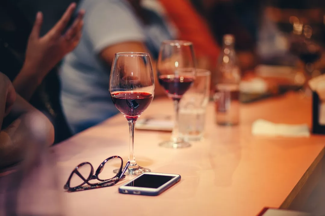 Ein Tisch mit zwei Gläsern Rotwein, einem Smartphone, einer Brille und einem Glas Wasser; im Hintergrund sitzen Menschen, die die entspannte Atmosphäre Mallorcas heraufbeschwören.