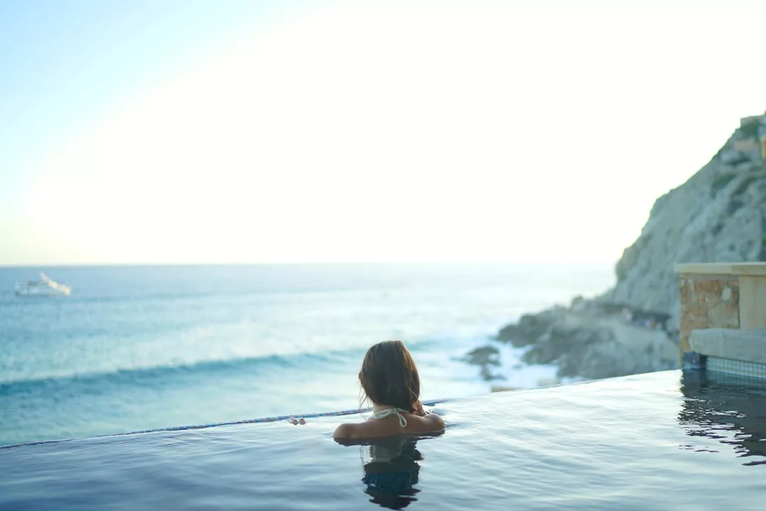 Eine Person mit langen Haaren entspannt sich in einem Infinity-Pool mit Blick auf das Meer auf Mallorca, mit Wellen und felsigen Klippen im Hintergrund unter einem strahlenden Himmel.