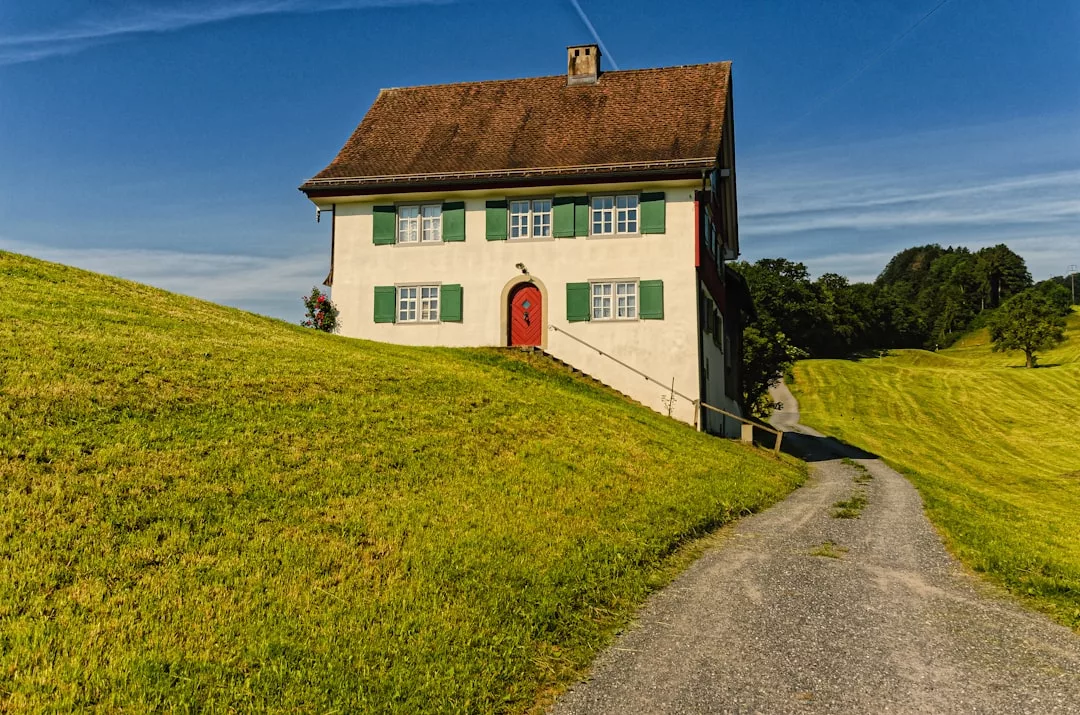Ein zweistöckiges Haus mit grünen Fensterläden und einer roten Tür steht auf einem grasbewachsenen Hügel neben einem Kiesweg unter dem klaren blauen Himmel Mallorcas.