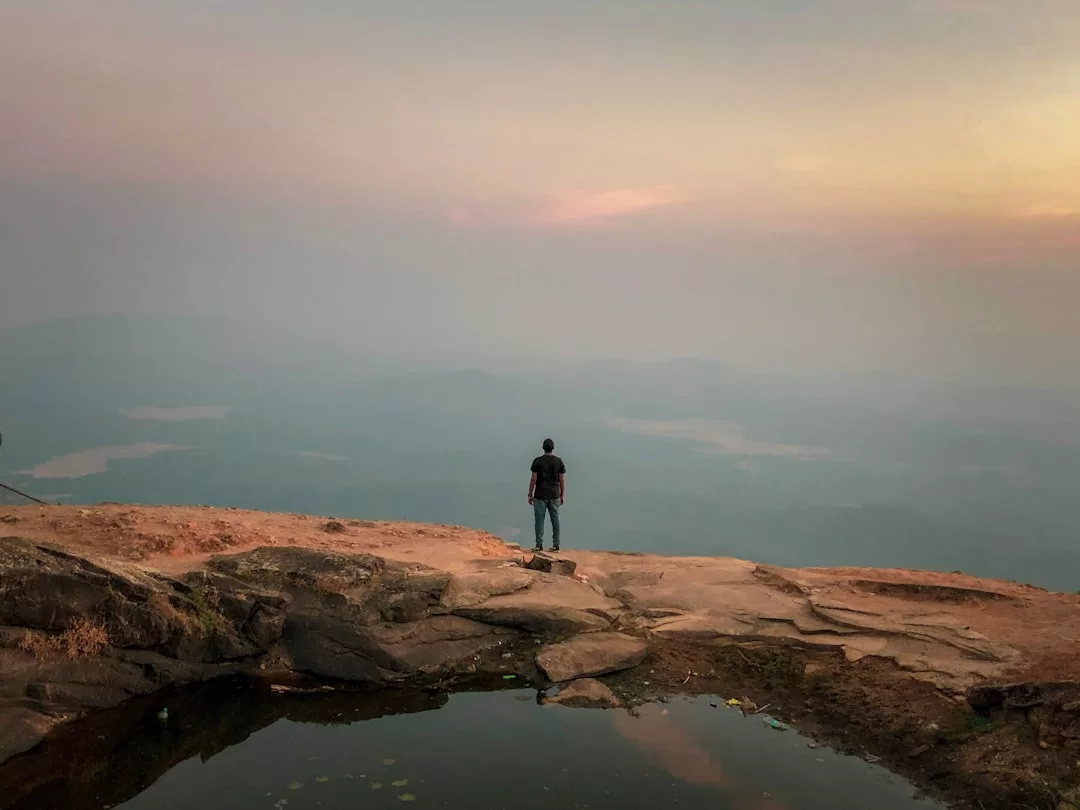 Eine Person steht auf felsigem Grund in der Nähe eines kleinen Wasserbeckens und blickt auf die dunstige Berglandschaft Mallorcas bei Sonnenuntergang oder Sonnenaufgang.