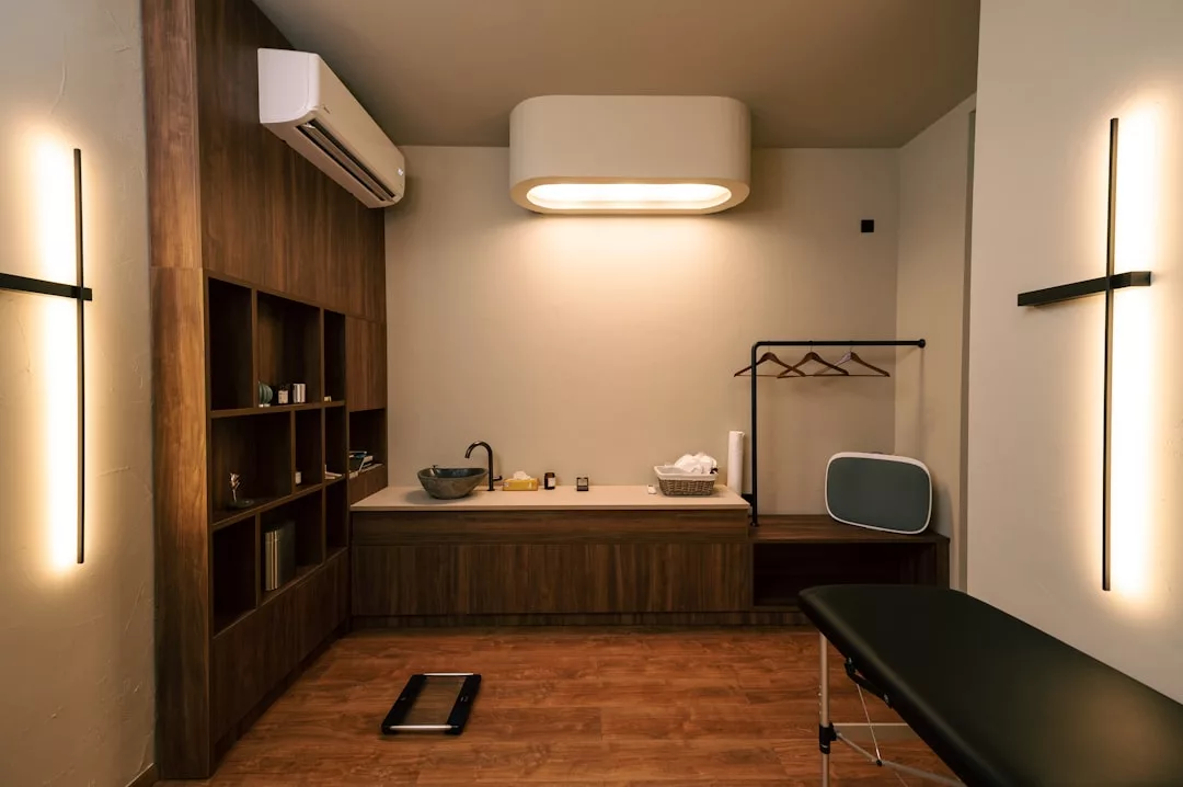 Modernes medizinisches oder Wellness-Untersuchungszimmer auf Mallorca mit Holzschränken, einem Waschbecken, Regalen, einem schwarzen Untersuchungstisch, einer Garderobe und einer warmen Raumbeleuchtung.